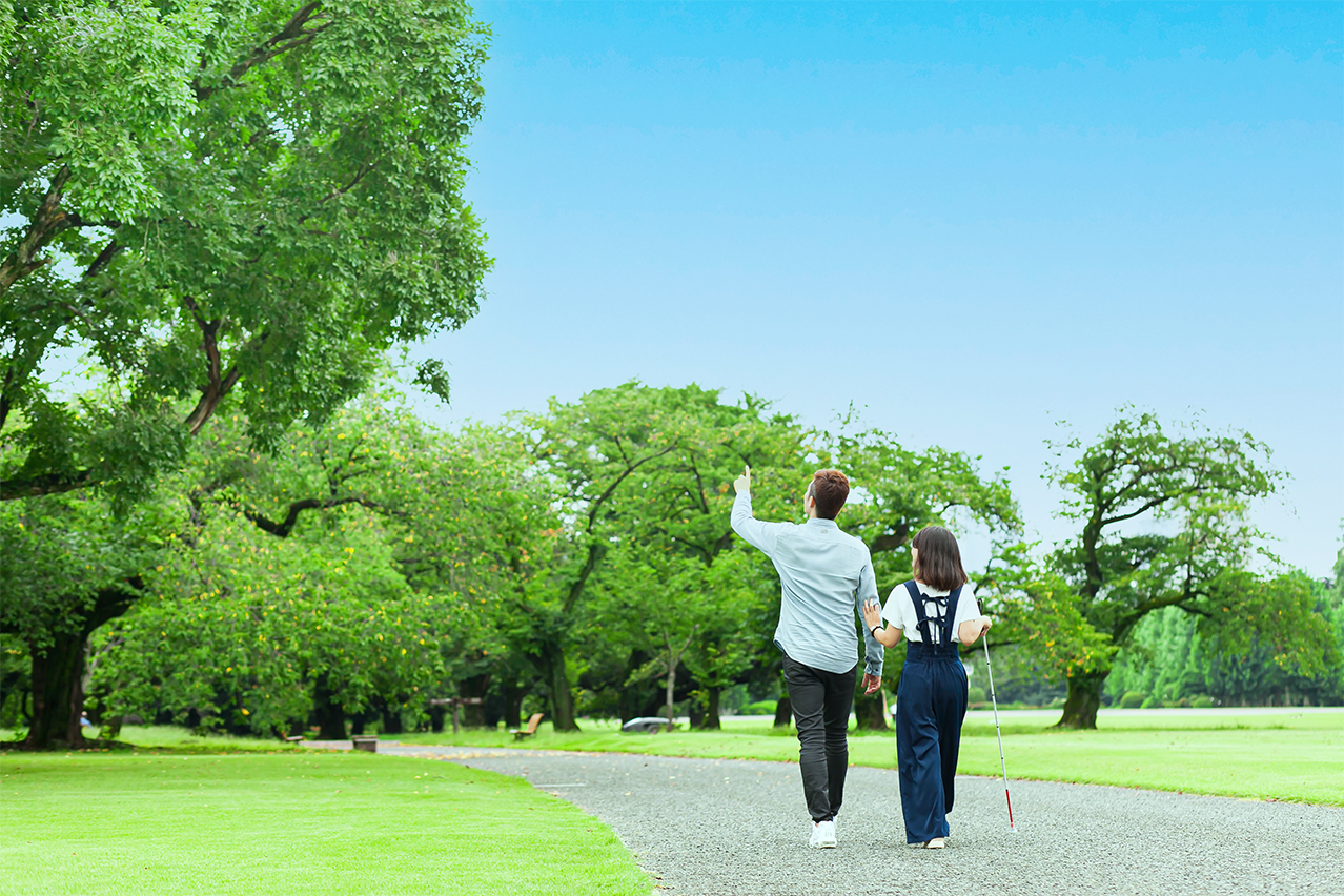青空のもと男性の腕につかまり一緒に散歩をする白杖の女性